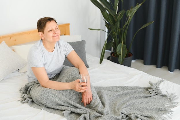 Blonde vrouw van middelbare leeftijd glimlachend gelukkig glas water liggend op het bed thuis