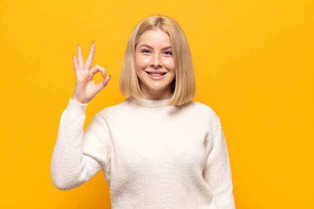 Blonde vrouw voelt zich gelukkig, ontspannen en tevreden, toont goedkeuring met ok gebaar, glimlachend