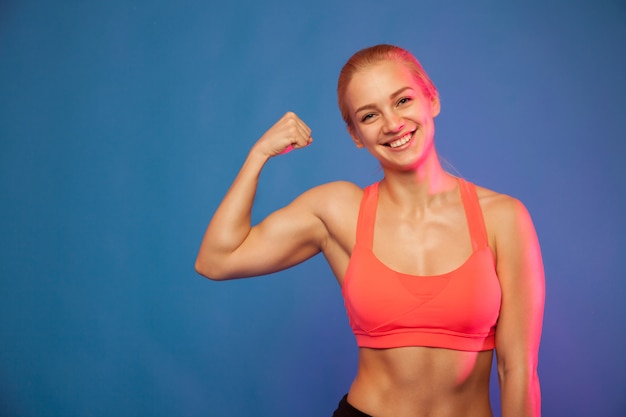 Blonde vrouwelijke atleet die bicepsen op blauwe achtergrond toont,