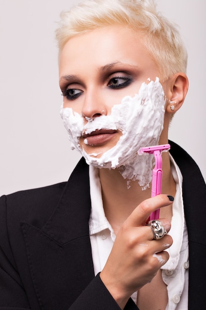 Blonde with a short haircut in a black jacket with shaving foam on her face. Pink women's razor.