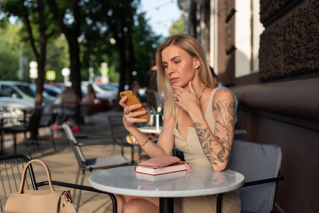 Una bionda con un tatuaggio sul braccio siede in un caffè estivo e parla al telefono con emozione