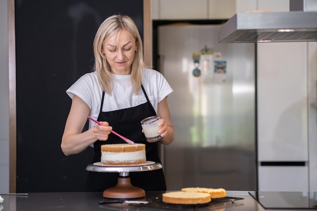 Blonde woman in black apron smearing fresh cake layers with sweet cream using pink brush