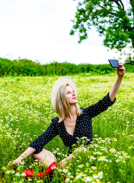 Блондинка в черном платье сидит на поле цветов ромашки и с помощью мобильного телефона