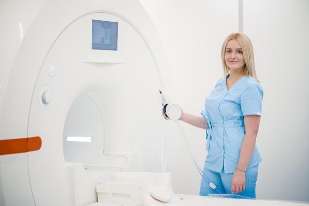 Blonde woman in blue uniform standing near MRI apparatus