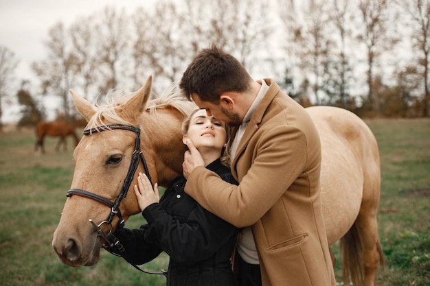 Blonde woman, brunette man standing in the field with brown horse. Woman wearing black clothes and man wearing brown coat. Man and woman hugging.