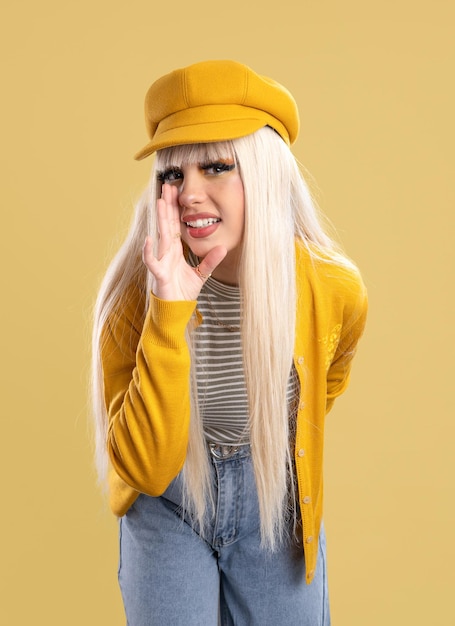 Blonde woman in cap and yellow jacket leaning towards the camera telling a secret, yellow background