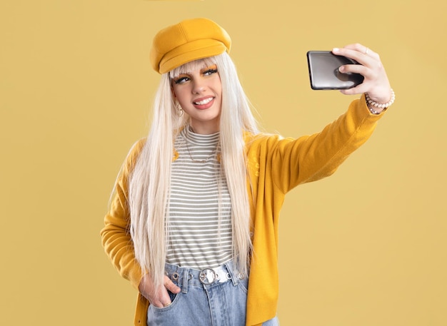 Donna bionda con berretto e giacca gialla che fa selfie con il telefono su sfondo giallo donna bionda con berretto e giacca gialla che scatta una foto con lo smartphone su sfondo giallo