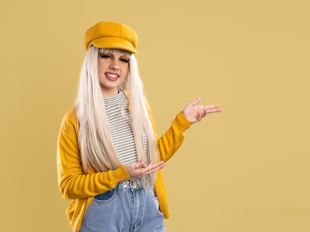 Blonde woman in cap and yellow jacket pointing fingers to the side over yellow background