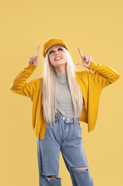 Blonde woman in cap and yellow jacket pointing fingers up on yellow background