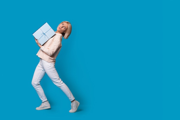 Donna bionda che porta una scatola con il regalo su uno studio di parete blu che fa pubblicità a qualcosa