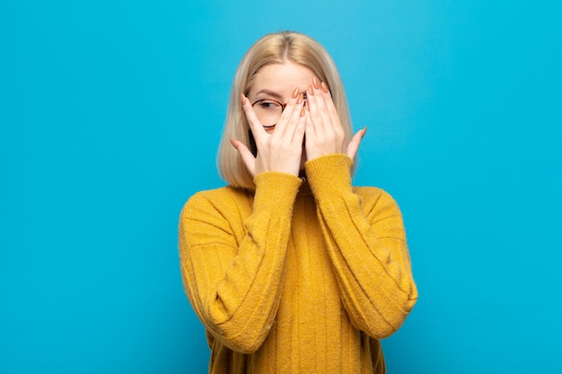 Blonde woman covering face with hands, peeking between fingers with surprised expression and looking to the side