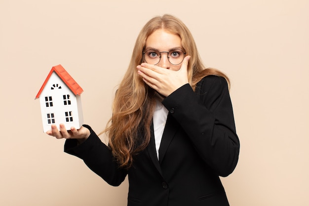 Blonde woman covering mouth with hands with a shocked, surprised expression, keeping a secret or saying oops