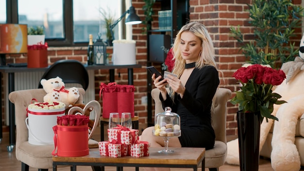 Blonde woman doing online shopping on smartphone with credit card to celebrate valentines day. Beautiful model enjoying love holiday surprise with romantic presents and flowers bouquets.