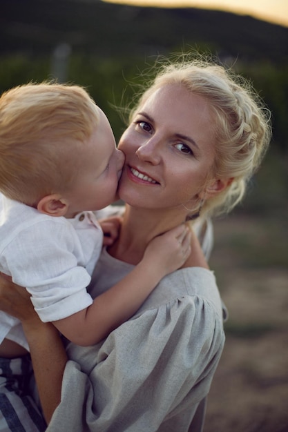 Photo blonde woman in a dress holds her baby boy in her arms in a vineyard during sunset