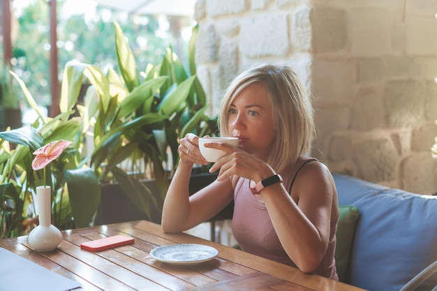 日当たりの良い屋外カフェに座って熱いお茶を飲むブロンドの女性