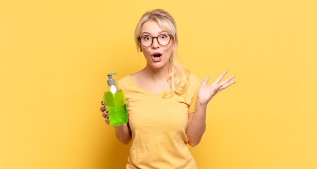 Blonde woman feeling extremely shocked and surprised, holding a soap bottle