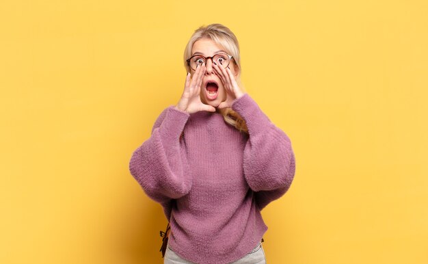 Blonde woman feeling happy, excited and positive, giving a big shout out with hands next to mouth, calling out