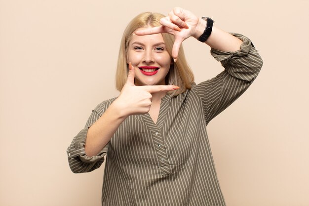 Blonde woman feeling happy, friendly and positive, smiling and making a portrait or photo frame with hands