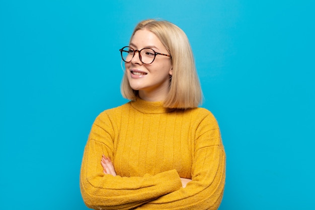 Blonde woman feeling happy, proud and hopeful, wondering or thinking, looking up to copy space with crossed arms