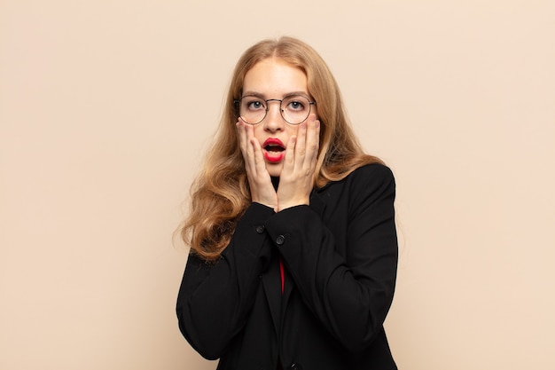 Blonde woman feeling shocked and scared, looking terrified with open mouth and hands on cheeks