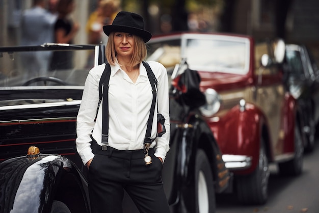 Blonde woman in hat and in black retro clothes near old vintage classic car.