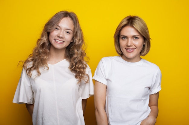 Blonde woman and her curly sister are smiling at camera on a yellow studio wall in the same clothes