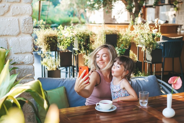 Blonde woman and her little daughter using smartphone for taking selfie in the cafe