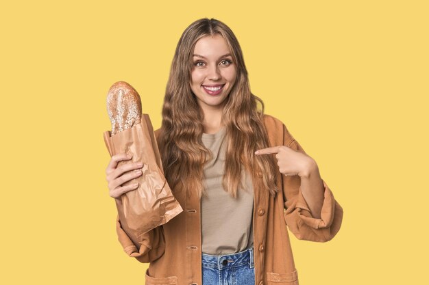 Блондинка, держащая хлеб, указывая рукой на рубашку, копирует пространство с гордостью и уверенностью.