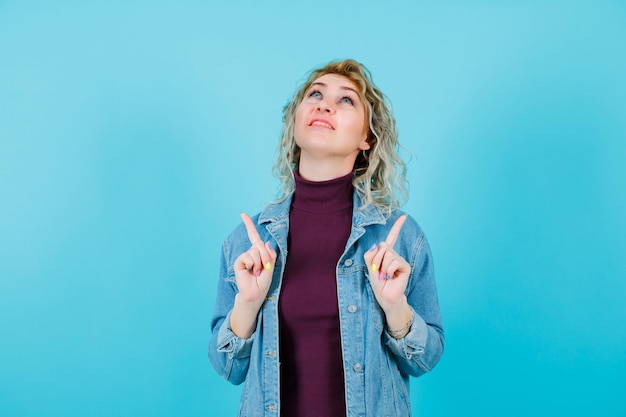 Blonde woman is looking and pointing up with forefingers on blue background