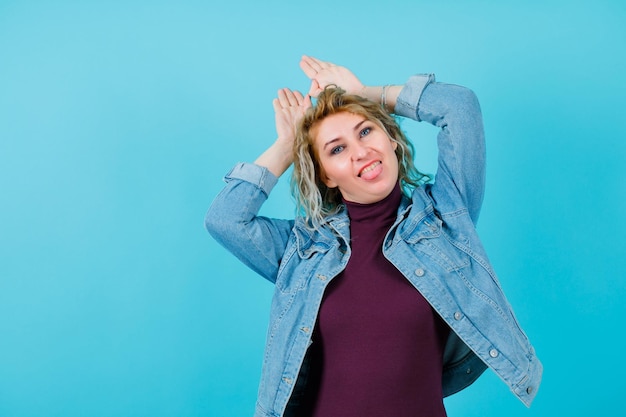 Blonde woman is showing rabbit gesture by holding hands on head on blue background