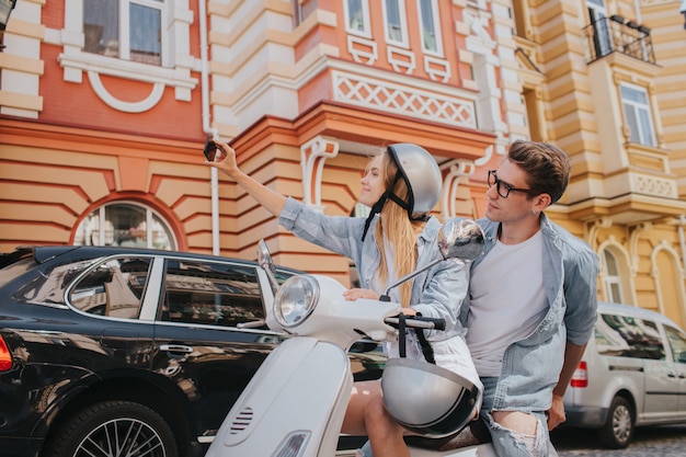 Blonde woman is sitting on bike with her boyfriend and taking selfie