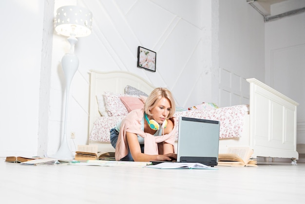 A blonde woman is sitting near the bed with a laptop and headphones
