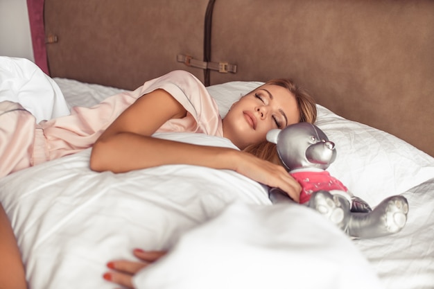 A blonde woman is sleeping with a silver bear in bed. Good Morning. Sleep and rest 