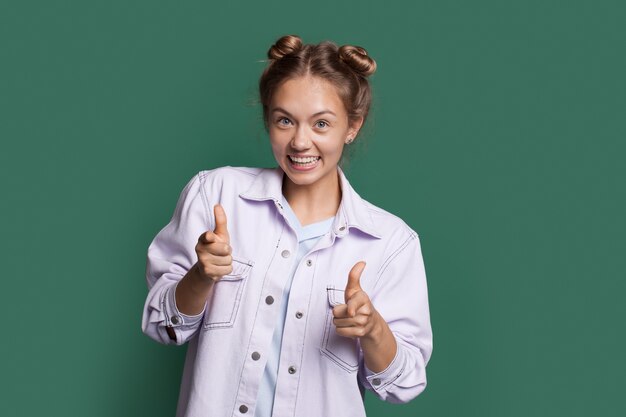 Blonde woman is smiling and pointing at camera wearing jeans clothes on a green studio wall