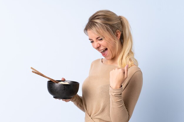 Blonde woman over isolated blue celebrating a victory while holding a bowl of noodles with chopsticks