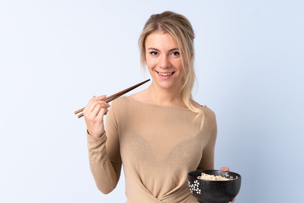 Blonde woman over isolated blue holding a bowl of noodles with chopsticks