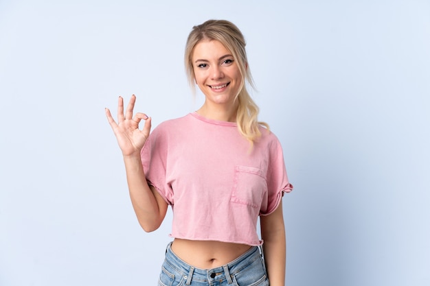 Blonde woman over isolated blue showing ok sign with fingers