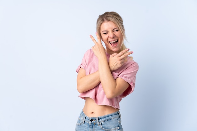 Blonde woman over isolated blue smiling and showing victory sign