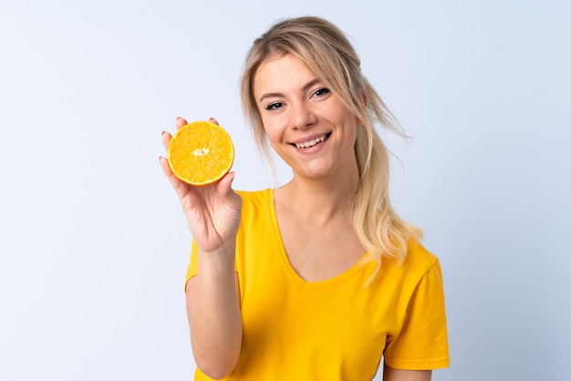 Blonde woman over isolated blue wall holding an orange