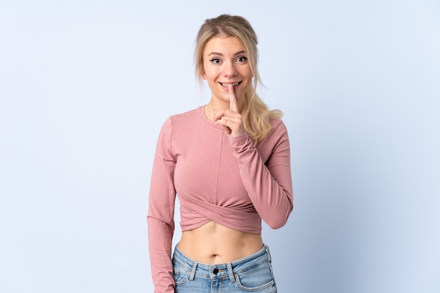 Blonde woman over isolated blue wall showing a sign of silence gesture putting finger in mouth