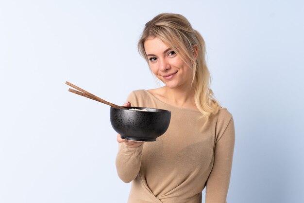 Blonde woman over isolated blue  with happy expression while holding a bowl of noodles with chopsticks