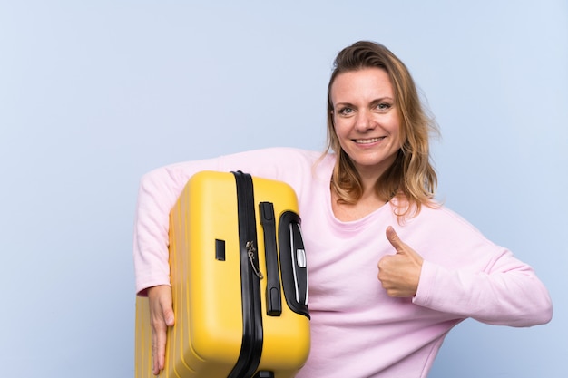 Blonde woman over isolated wall in vacation with travel suitcase and with thumb up