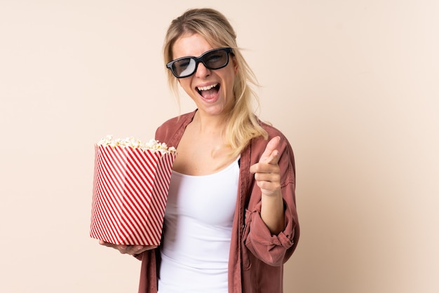 Blonde woman over isolated wall with 3d glasses and holding a big bucket of popcorns while pointing front