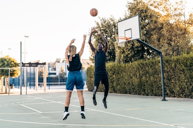 Blonde woman jumping as shooting a basketball at the basket\
while a man defends it outdoors