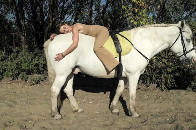 Блондинка в светлой одежде отдыхает на белой лошади.