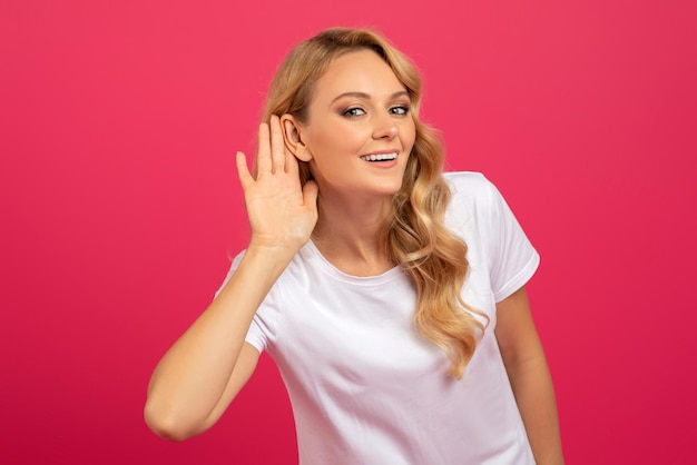 Blonde woman listening holding hand near ear over pink background