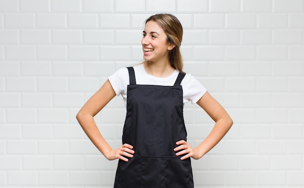 Foto donna bionda che sembra felice, allegra e sicura di sé, sorridendo con orgoglio e guardando a lato con entrambe le mani sui fianchi