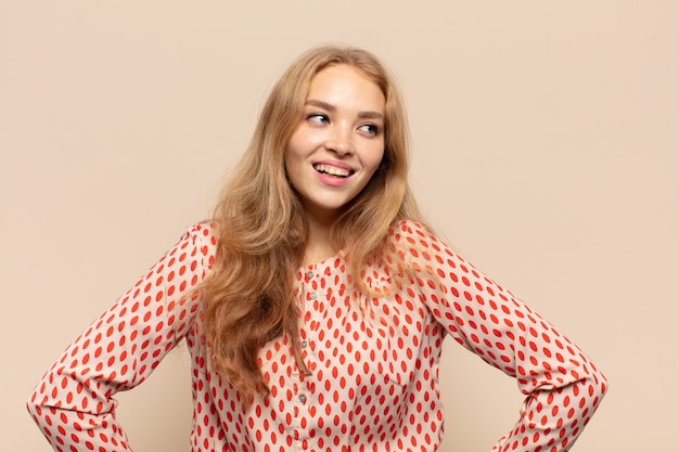 blonde woman looking happy, cheerful and confident, smiling proudly and looking to side with both hands on hips