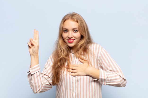 blonde woman looking happy, confident and trustworthy, smiling and showing victory sign, with a positive attitude