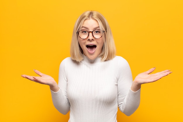 blonde woman looking happy and excited, shocked with an unexpected surprise with both hands open next to face